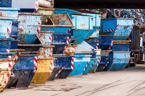 A row of large metal skip containers located outdoors, lined up along a paved area typically used for rubbish disposal or waste collection. The skips are made of heavy-duty steel with reinforced edges and are painted in various shades of blue, with some showing signs of rust, wear, and chipping paint that reveal a brownish corrosion underneath. The front sides of the skips feature diagonal reflective safety stripes in red and white, enhancing visibility. The containers are positioned on a concrete surface with visible tire marks and slight stains, suggesting frequent use. In the background, a dark waste collection truck with black and metallic elements is partially visible, indicating an active waste management operation. The scene is set during daylight with natural lighting, capturing the industrial environment associated with commercial or municipal rubbish removal services, reminiscent of facilities operated by companies such as Garden Clearance Harrow serving the Harrow area within Greater London.