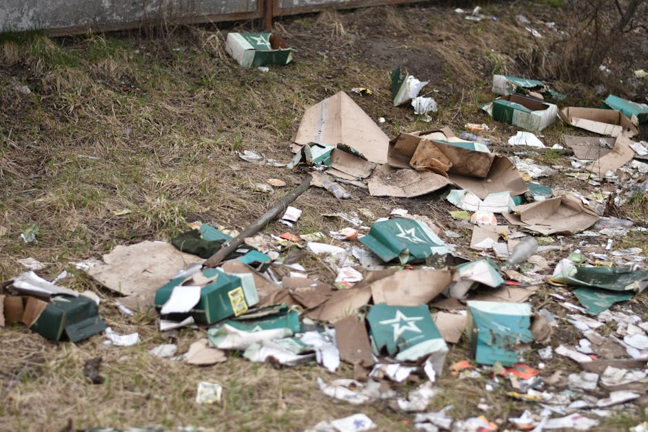 A row of four outdoor wooden rubbish bins with sloped, overhanging roofs, situated on a grassy area beside a sloped, wooded background. The bins are weathered, dark brown in colour, and evenly spaced