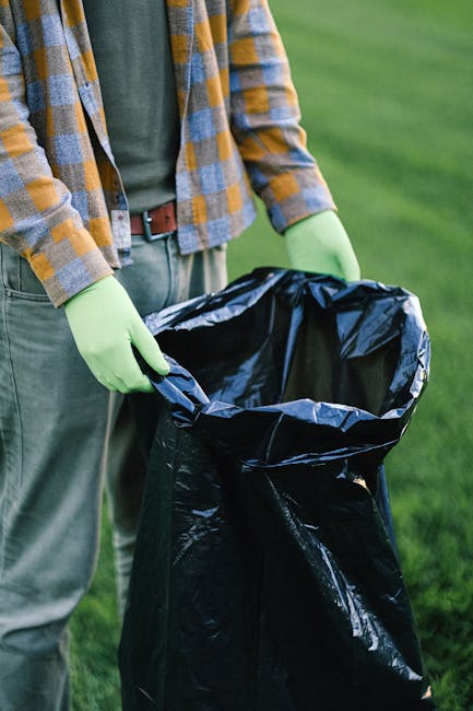A man with dark skin, wearing a black cap, red t-shirt, and light blue denim shorts, is standing on a sandy beach, holding a white rubbish bag in his left hand. Next to him, a young boy in a grey t-sh