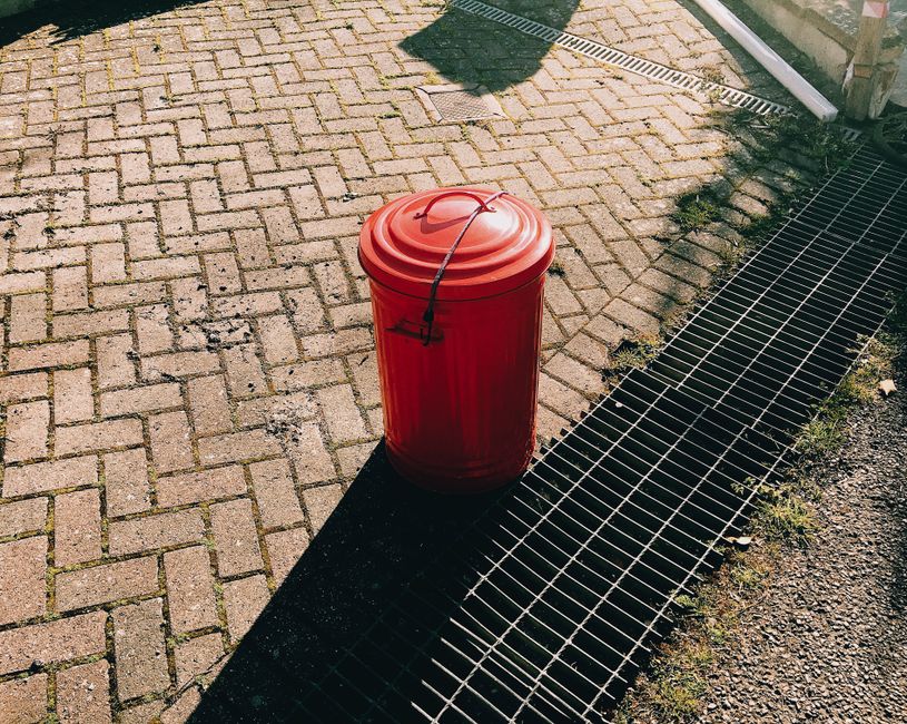 Two green wheeled rubbish bins with attached lids are positioned side by side on a concrete sidewalk close to the edge of a paved street. The bins are made of durable plastic with a smooth finish and 